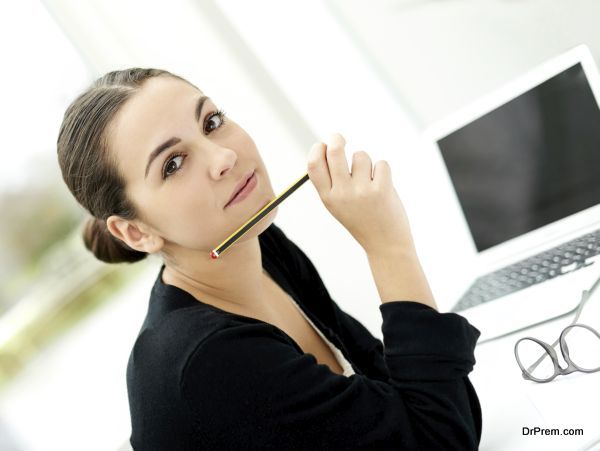Attractive stylish young businesswoman sitting at her desk with a laptop computer and her pencil raised to her chin looking at the camera with a serious expression