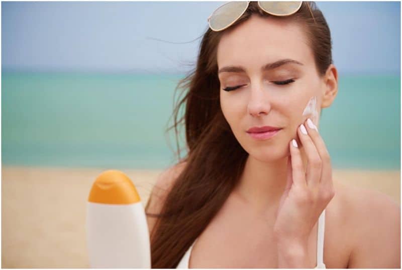 Woman holding sunscreen bottle applying sunscreen on her face at the beach