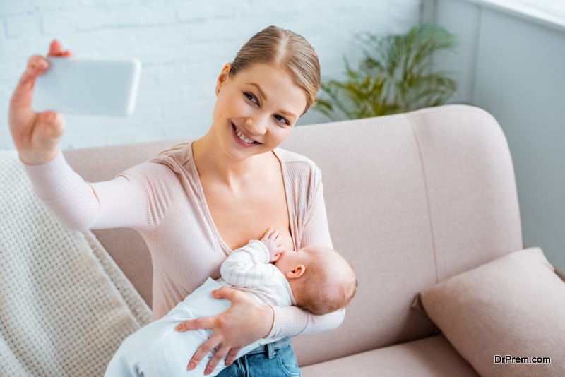 happy young woman breastfeeding baby and taking selfie with smartphone at home
