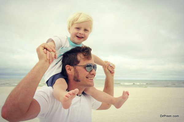 A young father has his toddler son on riding on his shoulders, and they are playing on the white sand beach by the ocean on a family summer vacation.