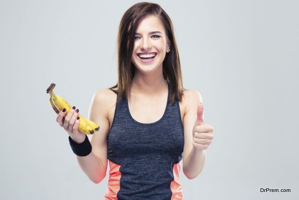 Laughing fitness woman holding banana and showing thumb up over gray background. Looking at camera