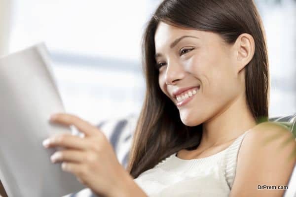 mixed race chinese asian / caucasian woman reading magazine relaxing sitting in sofa at home. Model smiling happy.