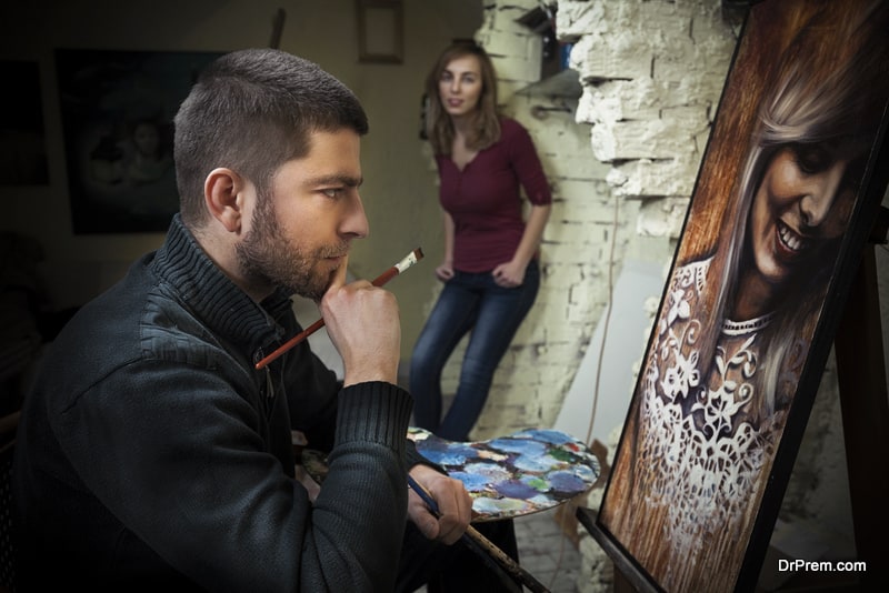 Young artist in his studio working on a portrait of beautiful young woman posing in the background