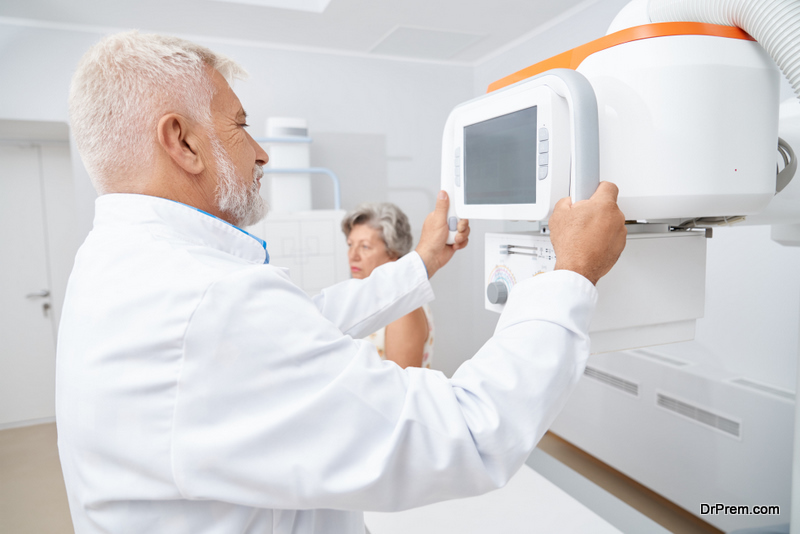 Old grey haired doctor using moving big ultrasound equipment looking at screen, preparing for procedure. Female patient waiting for usg checking of health, sitting in cabinet in modern hospital.