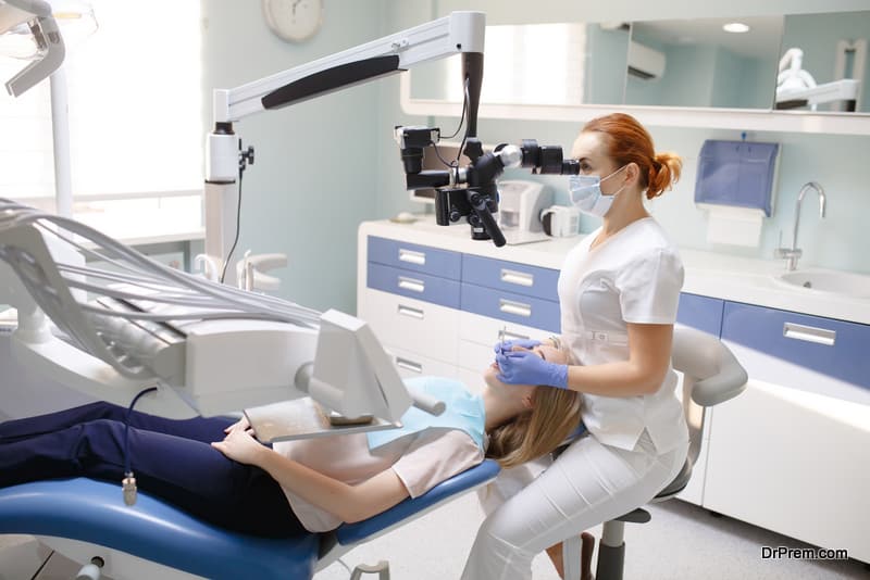 Female dentist with dental tools - microscope, mirror and probe treating patient teeth at dental clinic office.