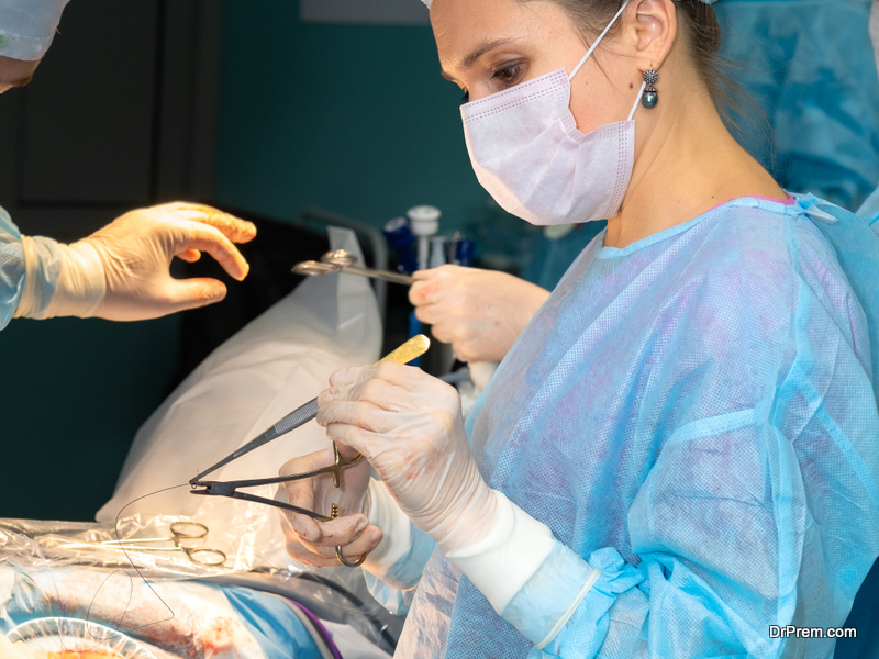 Female surgeon sutures the patient's skin during surgery. The doctor in sterile gloves holds the surgical thread with tweezers and forceps. Selective focus.
