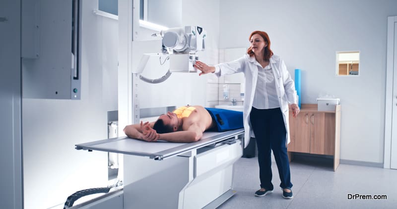 Mature woman in medical robe covering shirtless man with lead apron and turning on X ray machine then walking away on lab of contemporary hospital