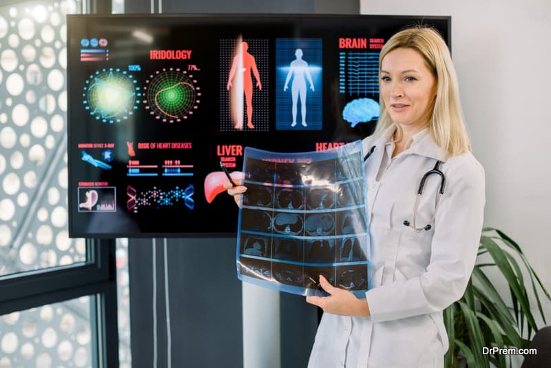 Medical conference, healthcare, computed tomography concept. Attractive middle aged blond female doctor radiologist, holding computed tomography x-ray image, standing in front of big digital screen.