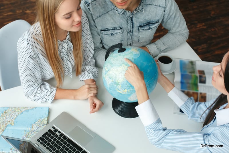 Young man and woman in a tour agency with a travel agent choosing destination on a globe looking curious smiling