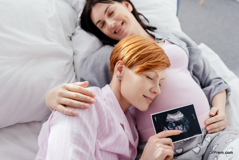 Smiling woman holding ultrasound scan of baby near belly of pregnant girlfriend on bed