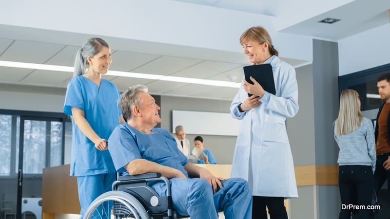 In the Hospital Lobby, Nurse Pushes Elderly Patient in the Wheelchair, Doctor Talks to Them while Using Tablet Computer. Clean, New Hospital with Professional Medical Personnel.