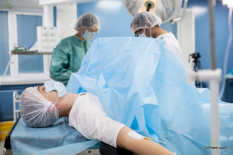 Young male patient in protective mask and headwear lying on operating table while two surgeons preparing medical instruments for operation