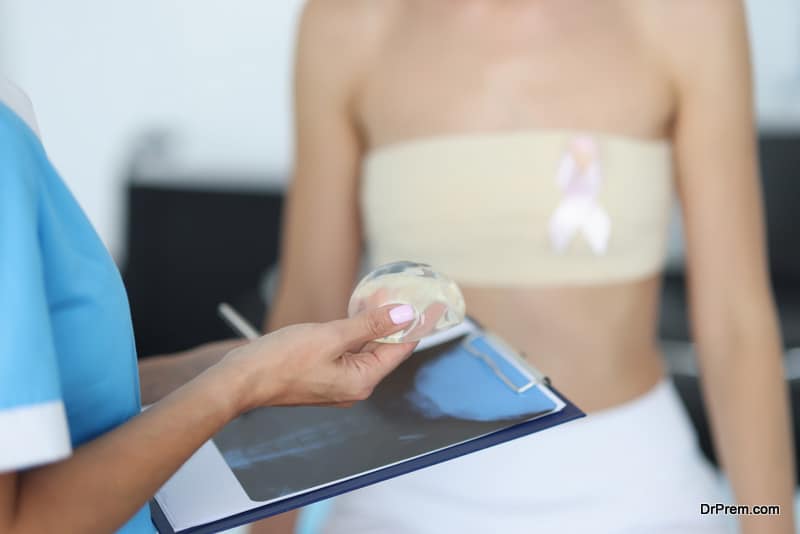 Doctor's hands are holding breast implant in front of clipboard with patient in background. Mammaplasty breast correction concept.