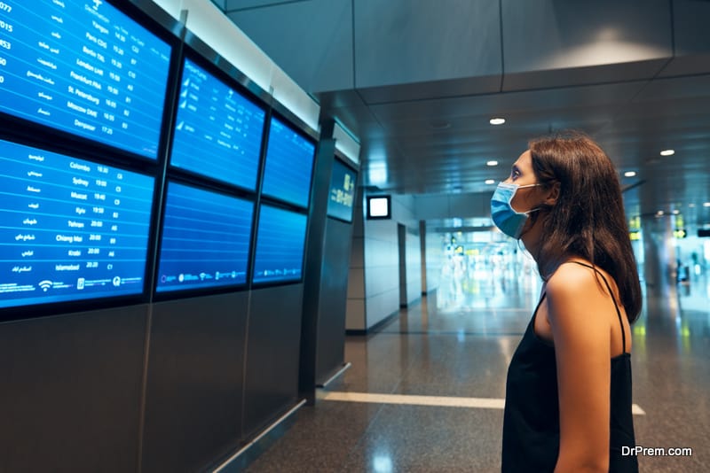 Woman in virus protection face mask looking at information board checking her flight in international airport. Departure board, flight status