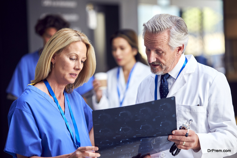 Two Doctors In White Coat And Scrubs Discussing Patient Scan In Busy Hospital