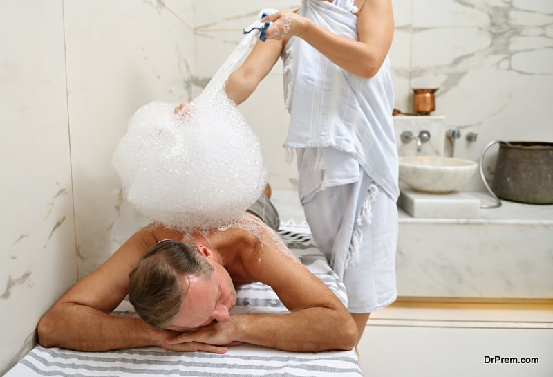 Handsome relaxed mature European man enjoying foam bath in Turkish hammam, lying on a marble surface while unrecognizable masseuse performing him a foam massage at wellness spa center
