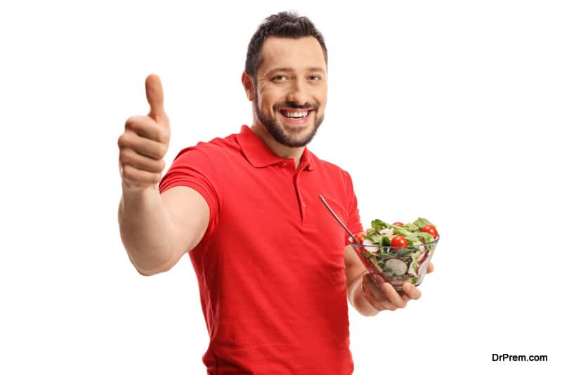 Smiling young man in a red t-shirt eating a healthy fresh salad and showing thumbs up isolated on white background