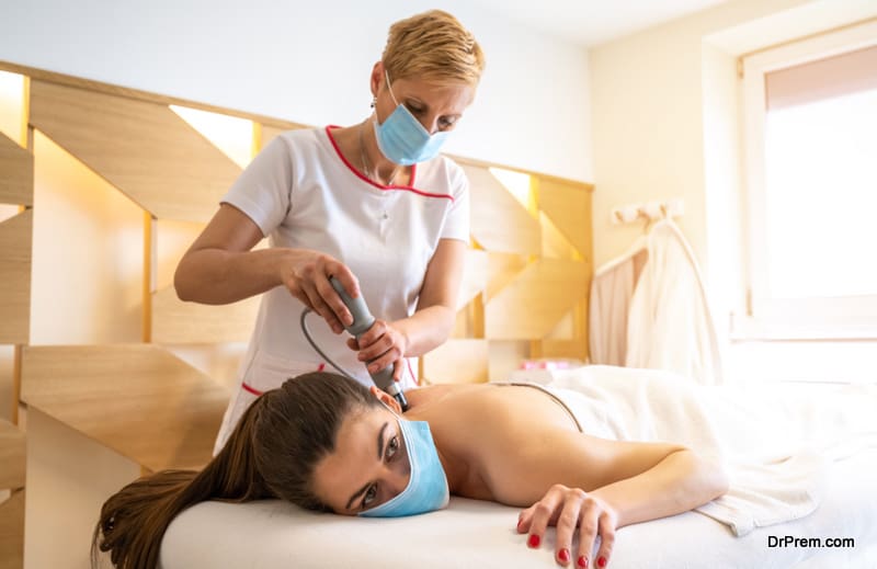 Extracorporeal shockwave therapy in beauty salon. Pain relief, normalization and regeneration,stimulation of healing process. Physical therapy for neck and back muscles,spine with shock waves. Young woman wearing protective mask during treatment in salon.