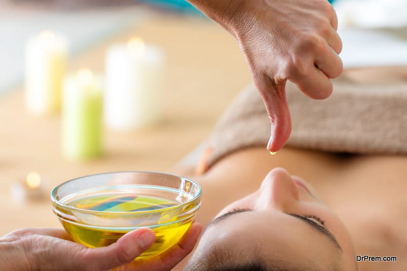 Macro close up of hand with oil drop above woman’s head in spa. Aromatic oil in glass bowl next to woman at Ayurvedic massage.