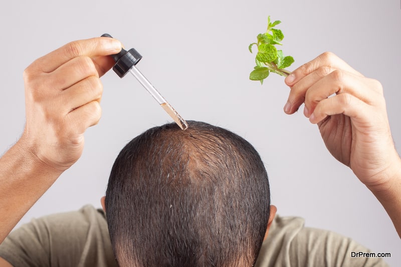 young guy showing mint leaves and apply hair essential oil in bald patches against grey background.