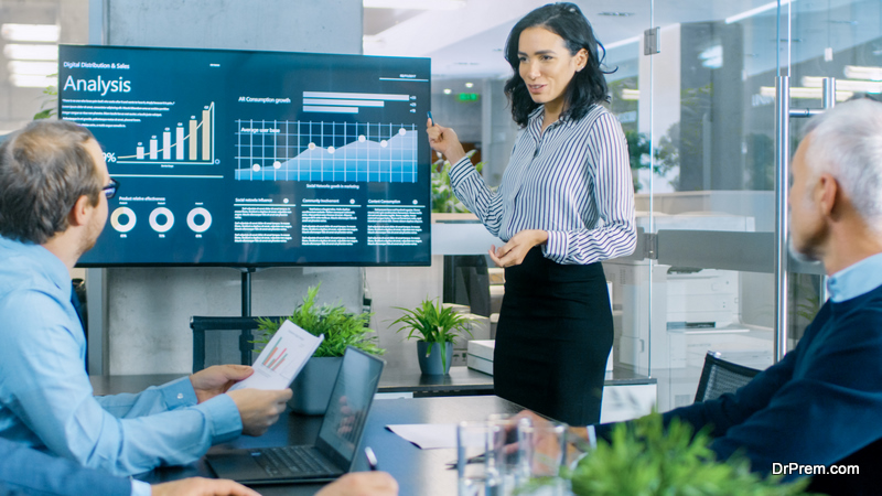 Beautiful Businesswoman Gives Report Presentation to Her Business Colleagues in the Conference Room, She Shows Graphics, Pie Charts and Company's Growth on the Wall TV.