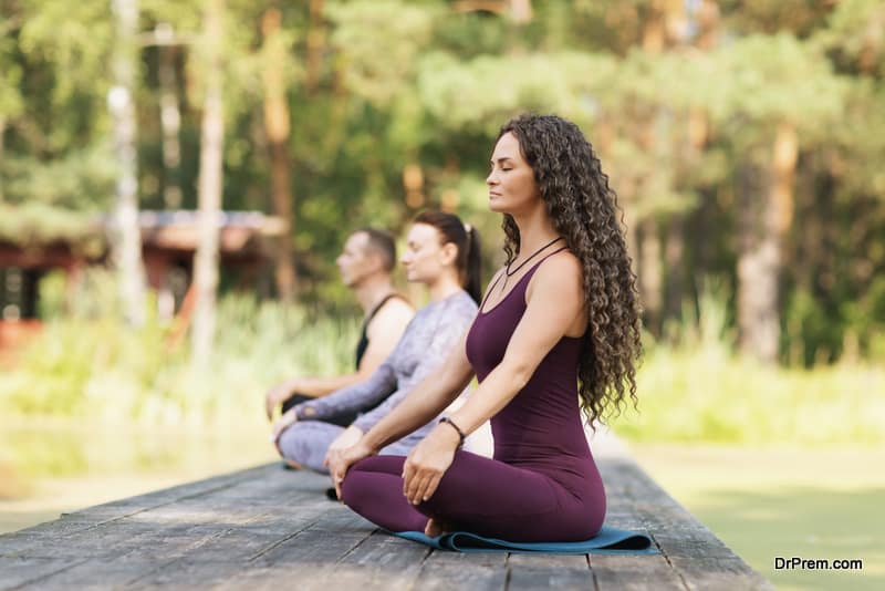 Beautiful woman trainer with two students sitting on a mat in a lotus position, are engaged in meditation on a wooden bridge in a park on a sunny summer morning