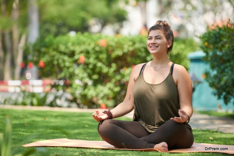 Beautiful young woman sitting in lotus position on mat in summer park