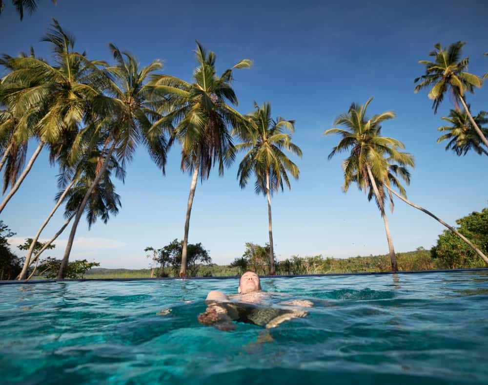 man-is-swimming-on-back-in-pool-against-palm-trees