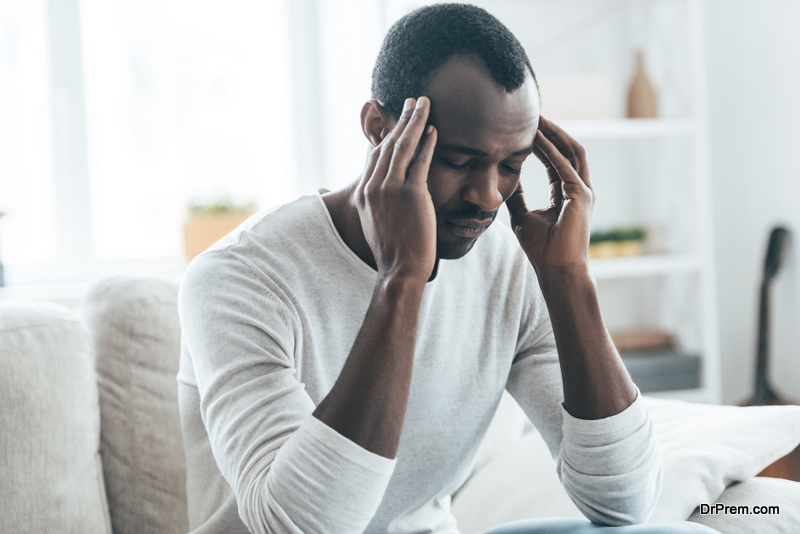 Young African man touching head with hands and keeping eyes closed while sitting on the sofa at home