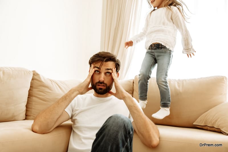 Dad is having a headache while his little daughter is jumping on the couch at home