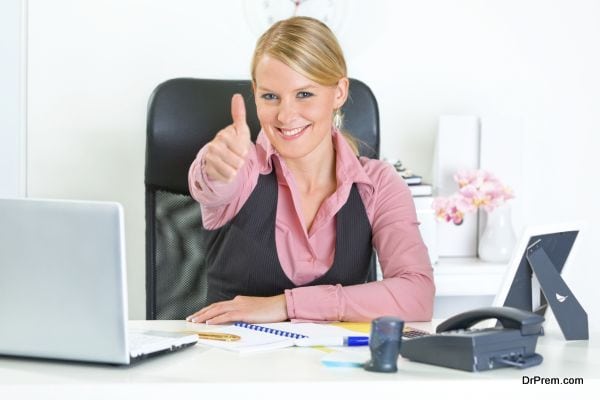 Smiling modern business woman sitting at office desk and  showing thumbs up gesture