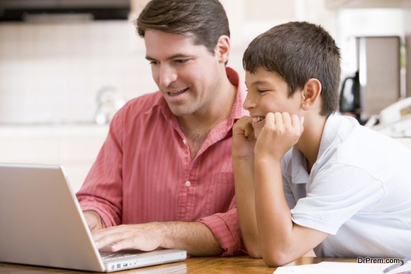 Man helping young boy in kitchen with laptop smiling